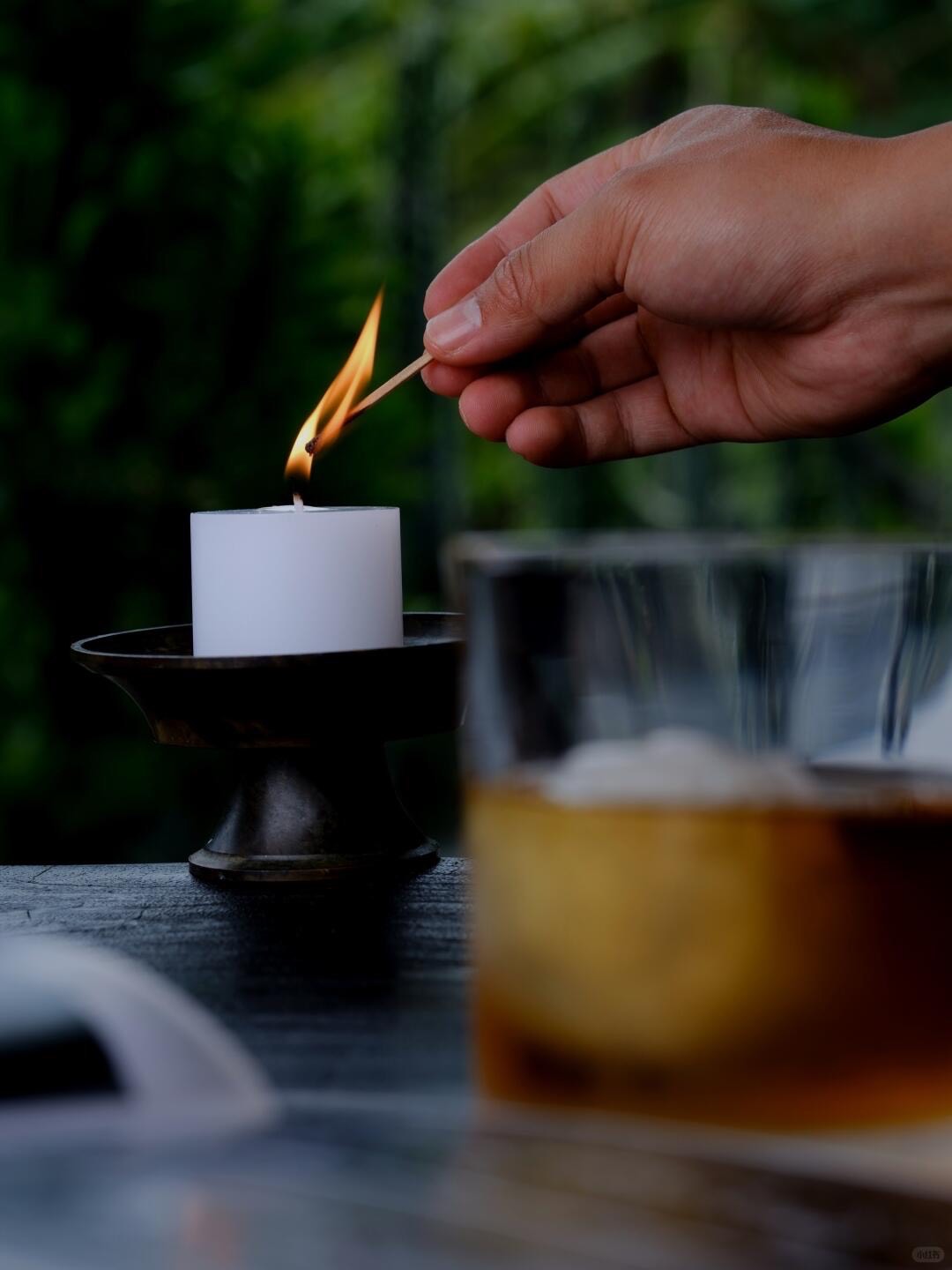 Hand lighting a match over a white pillar candle beside a glass of amber spirit on a dark table with greenery behind.