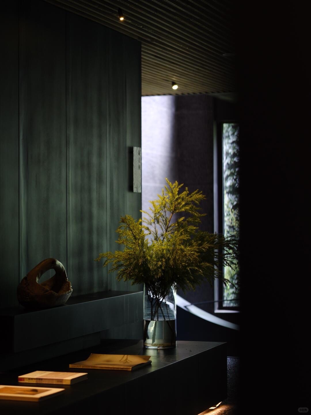 Dark green-panelled lobby corner with a glass vase of green foliage and books on a low bench.