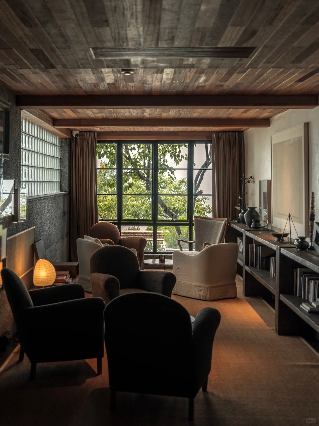 Warm timber-ceilinged lounge with clustered armchairs, book shelving and a tall window onto a leafy garden.