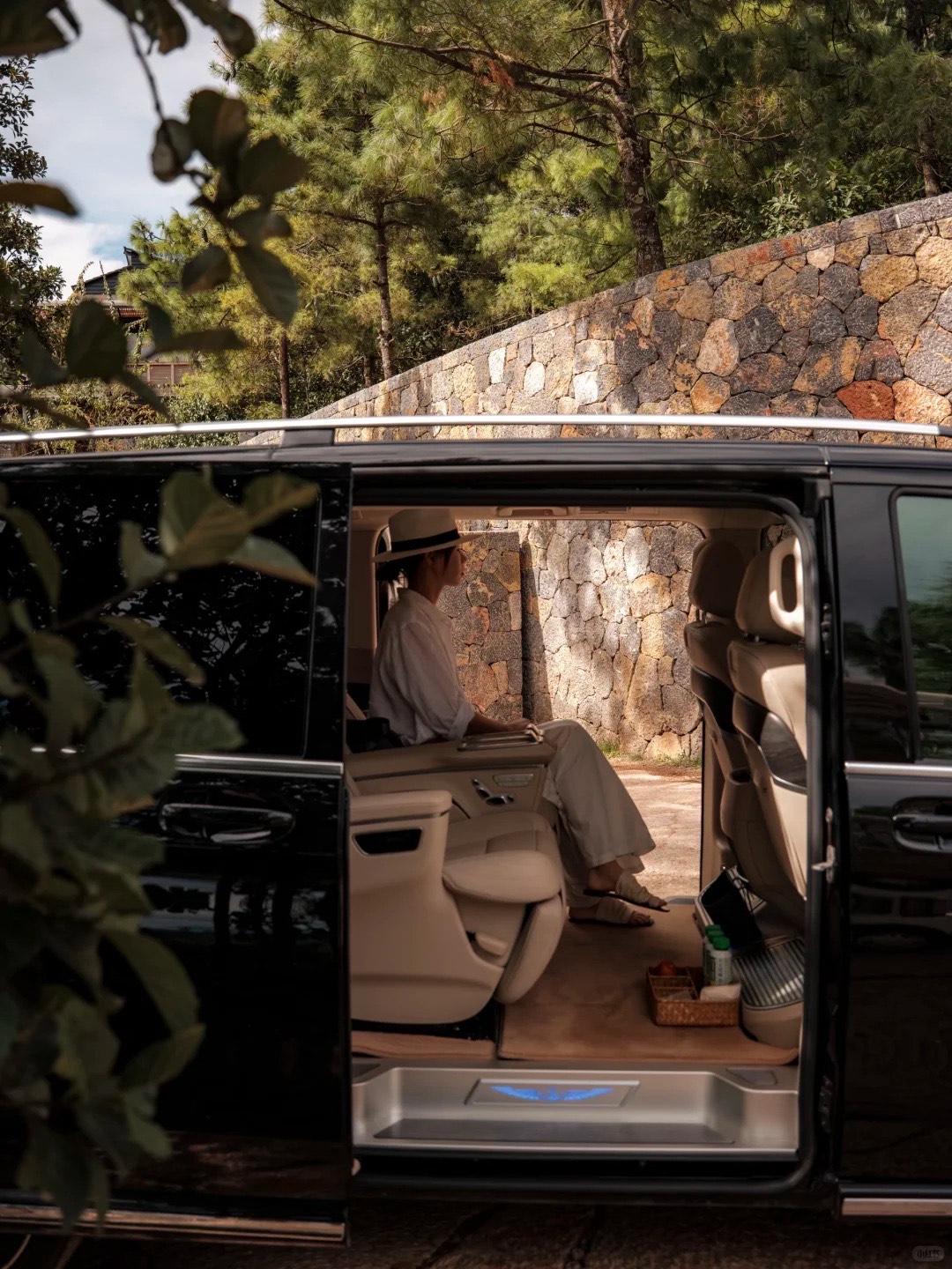 Guest in a sunhat seated in a chauffeured black luxury van with cream seats beside a curved dry-stone wall and pines.