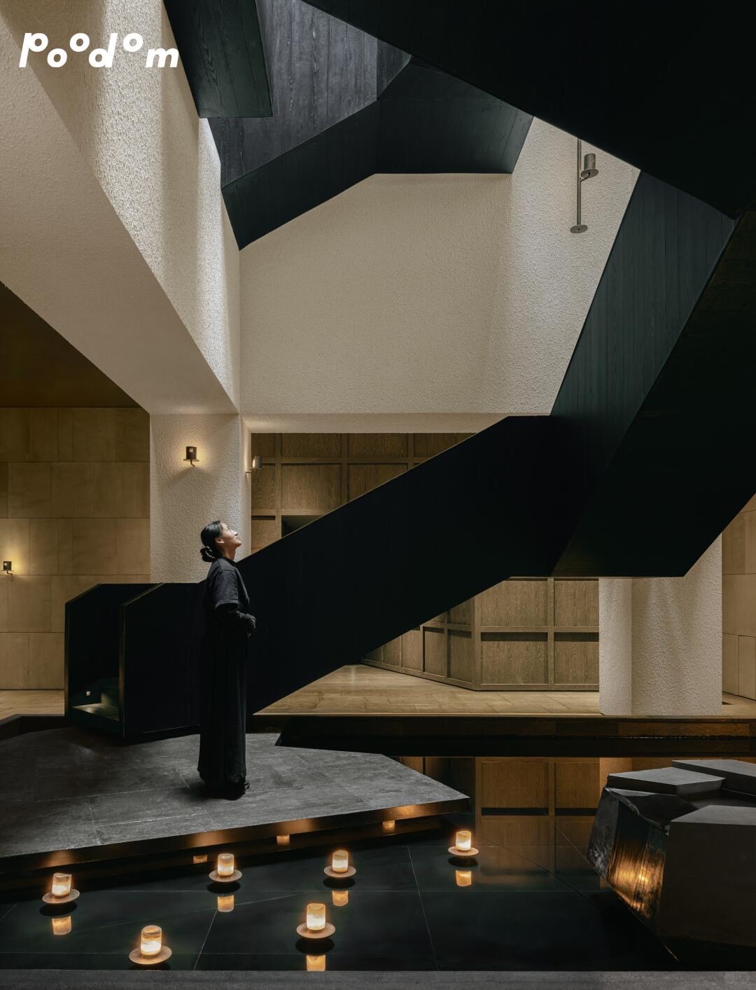 Woman in black standing by a candlelit reflecting pool beneath a dramatic black staircase in Poodom's soaring atrium.