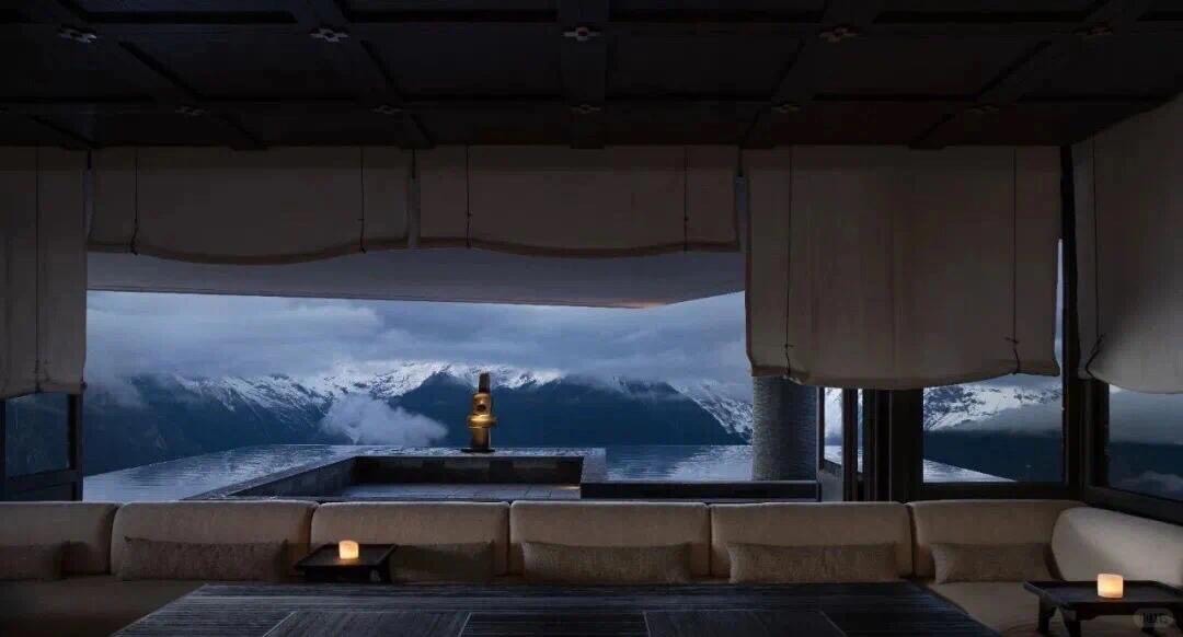 Low-lit lounge with soft sofas framing an infinity pool and snow-capped mountains wrapped in cloud at dusk.