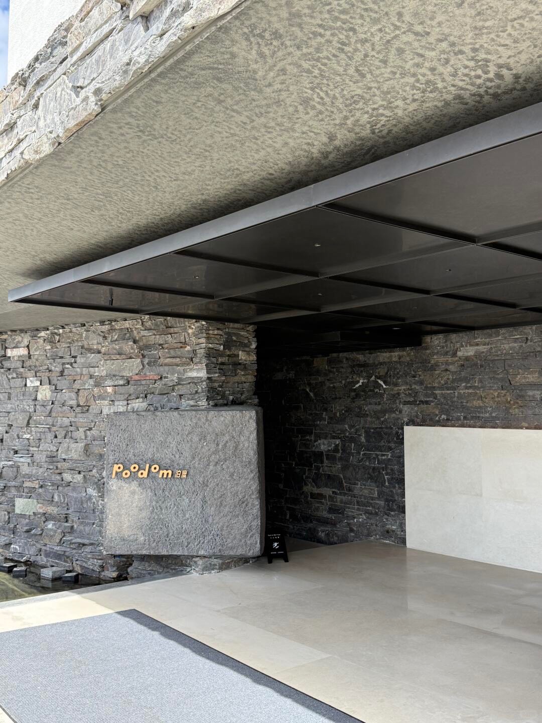 Poodom entrance framed by dark stacked-stone walls and a steel canopy over a polished stone-tiled arrival floor.