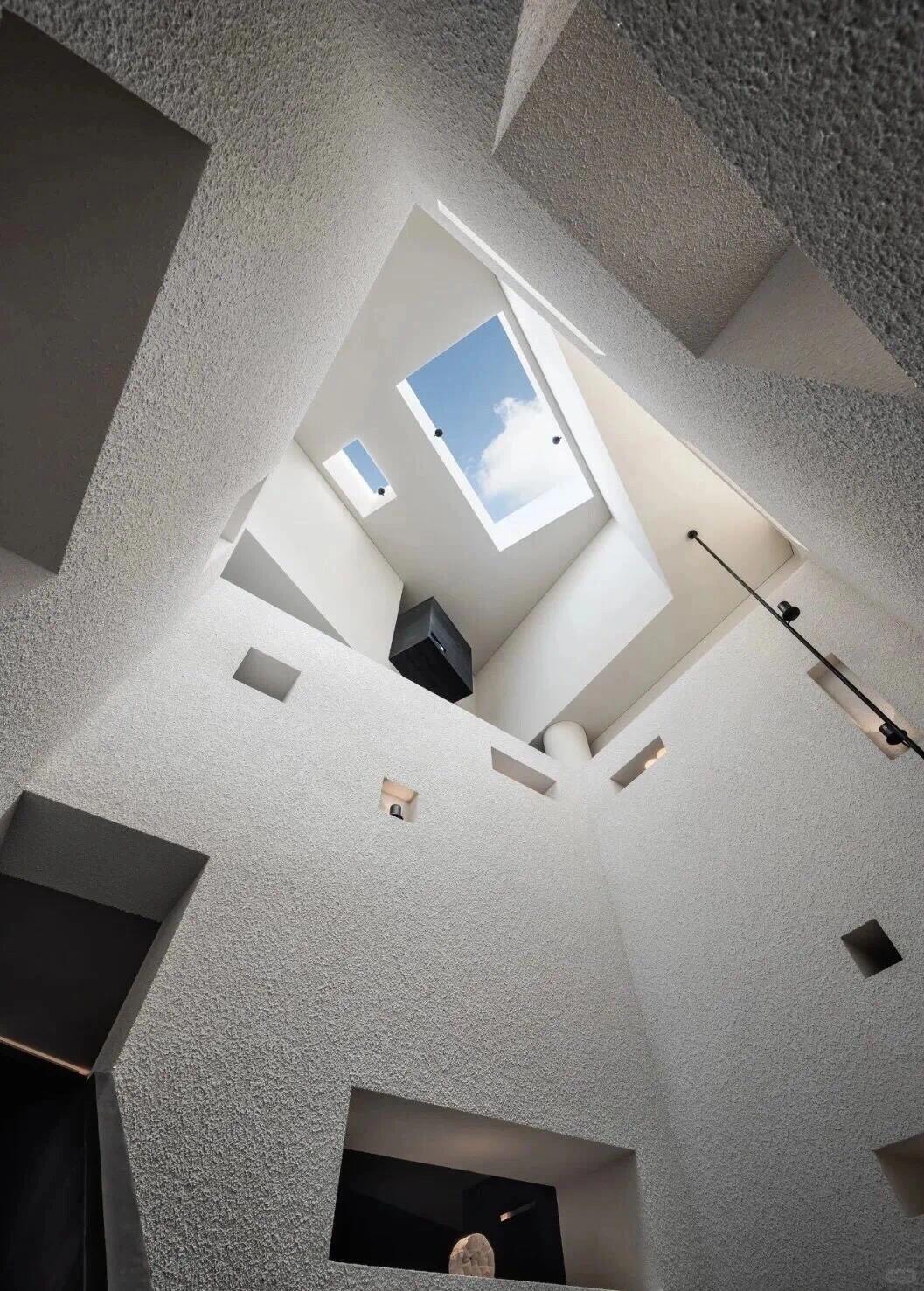 Looking up through a sculptural textured-concrete atrium to an angled skylight opening onto blue sky and cloud.