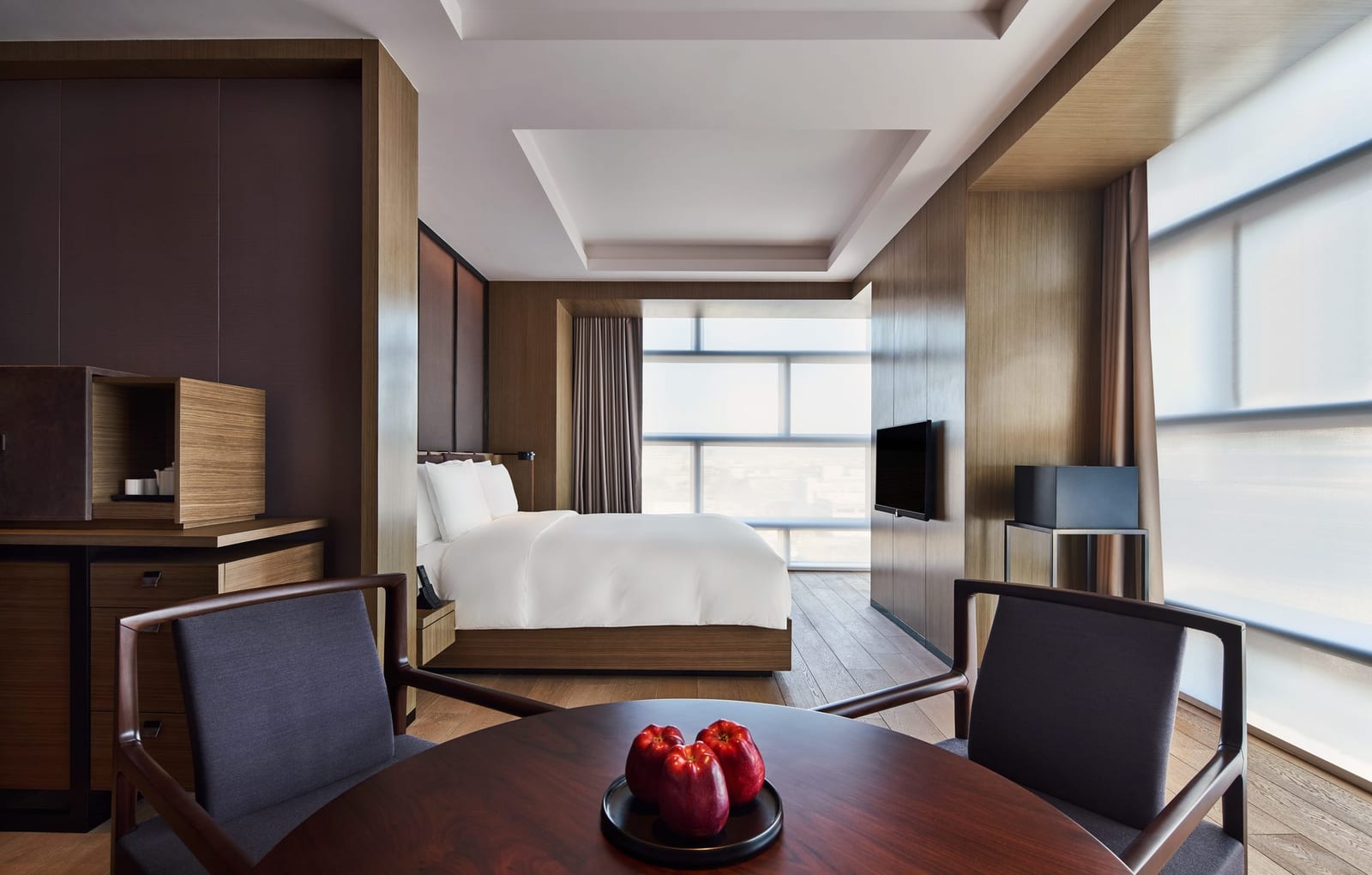 The PuXuan Hotel and Spa, Beijing — a timber-panelled guest room, a round table set with red apples in the foreground and a king bed before floor-to-ceiling windows