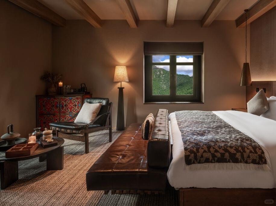 Earth-toned bedroom with white bed, patterned throw, leather bench and painted cabinet beside a forest-view window.