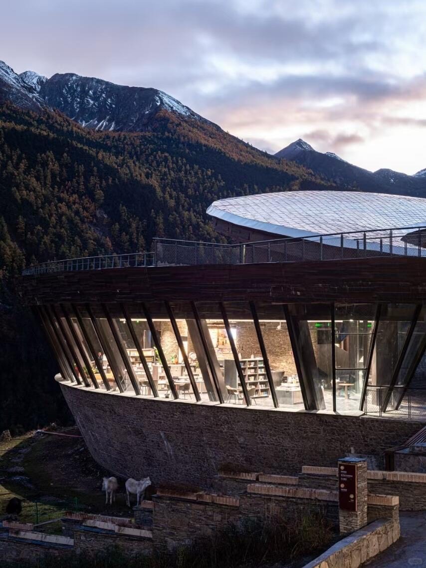 Curved glass-and-stone pavilion library set against forested mountains at dusk, with grazing cattle below.