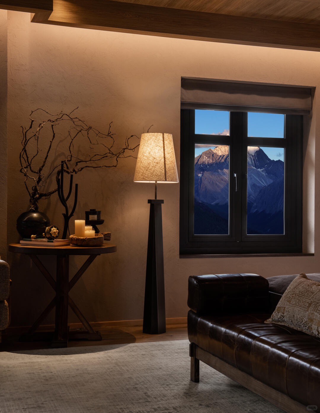 Warmly lit corner with floor lamp, leather sofa and console table beside a window onto snow-dusted peaks at dusk.