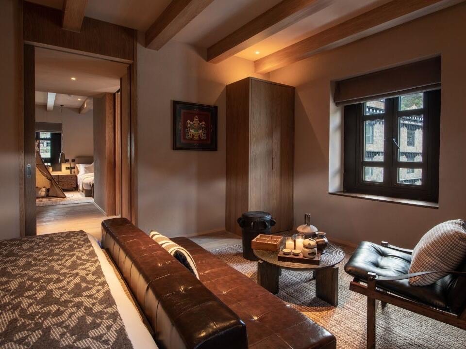 Warm-toned suite with leather bench, lounge chair and framed Tibetan artwork beside a window onto stone houses.