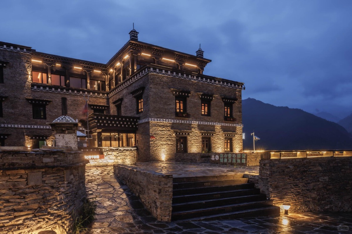 Illuminated Tibetan-style stone lodge with carved trim and mountains behind at blue-hour dusk.