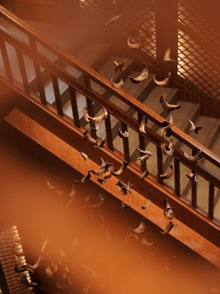 Suspended sculpture of hanging dried leaf forms above a warmly lit timber staircase and railing.