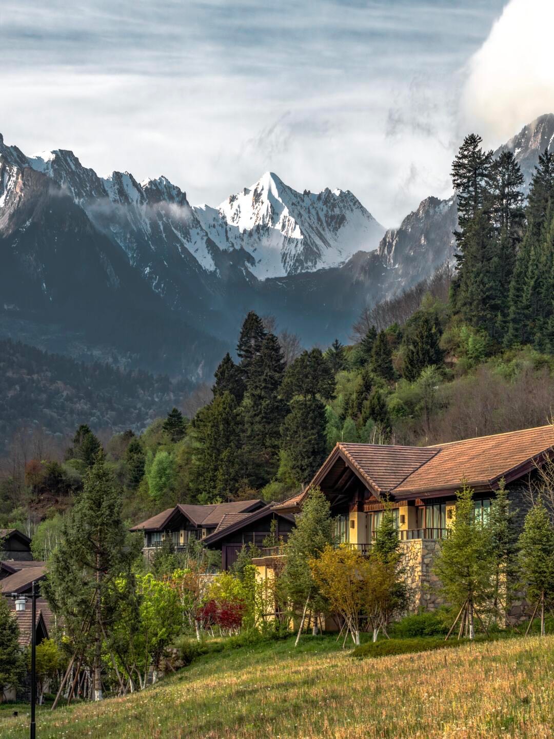 Timber-and-stone lodges set on a grassy hillside below forested slopes and snow-capped peaks.