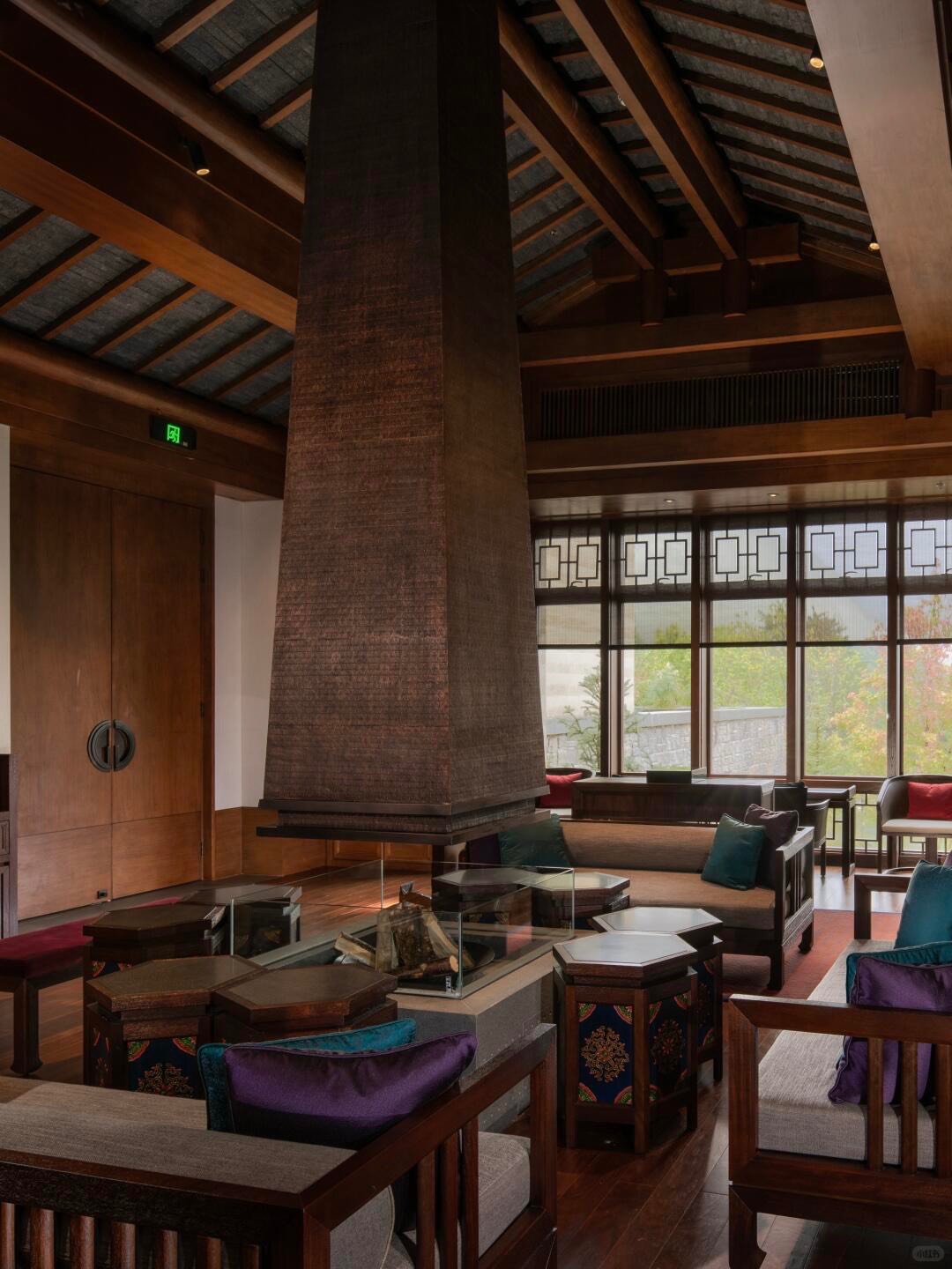 Lofty timber-beamed lounge with central chimney fireplace, low seating and lattice windows onto greenery.