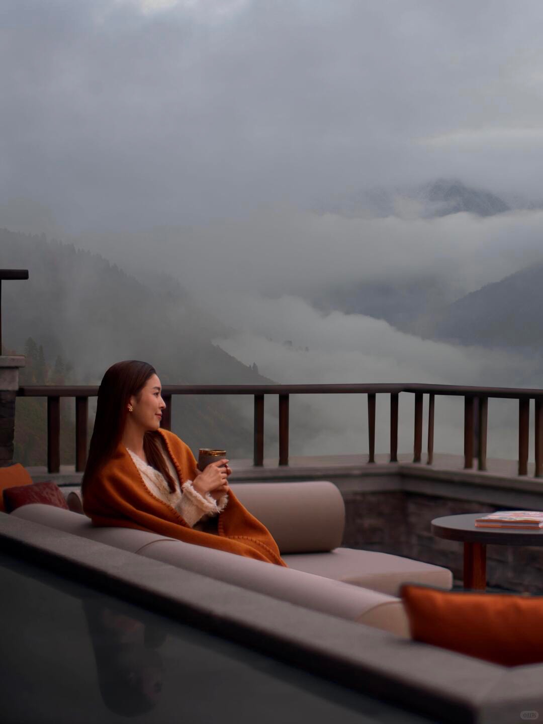 Guest wrapped in an orange blanket relaxing on a terrace sofa overlooking mist-shrouded forested mountains.