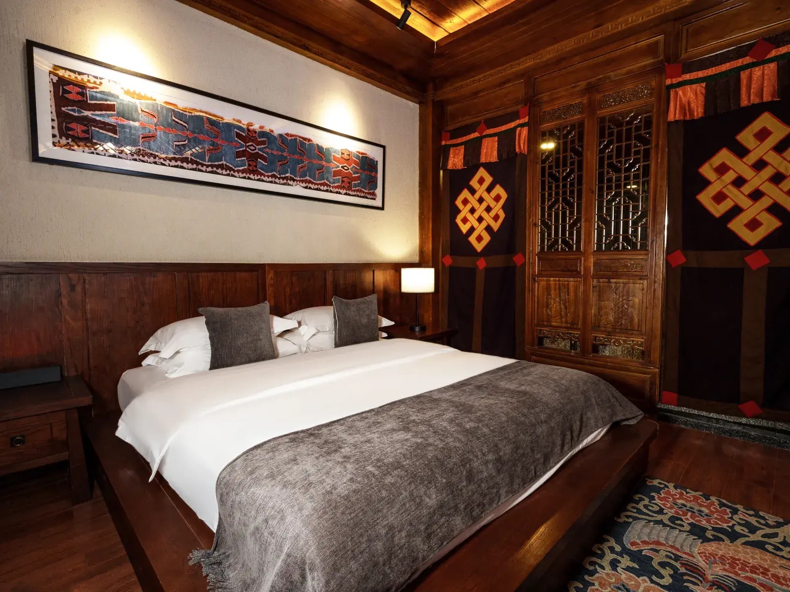 Sangzhu Bieyuan — a guest room panelled in dark carved wood, a woven Tibetan textile above the bed and endless-knot hangings flanking the lattice window