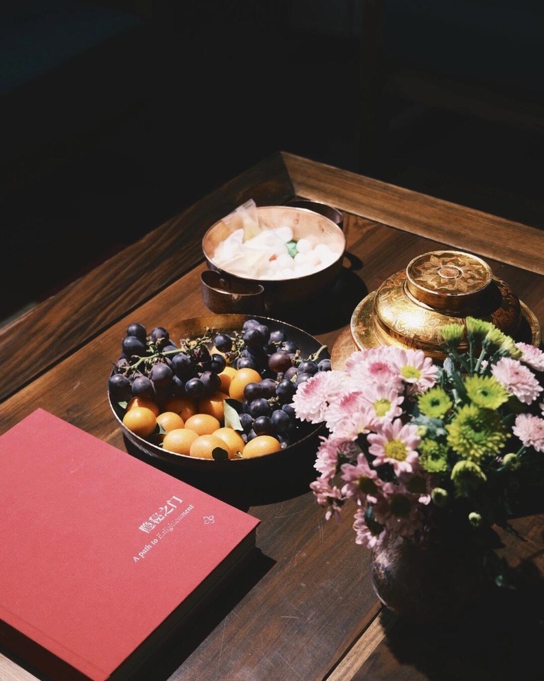 Wooden tray with fresh fruit, a brass lidded pot, chrysanthemums and a red welcome book on a table.