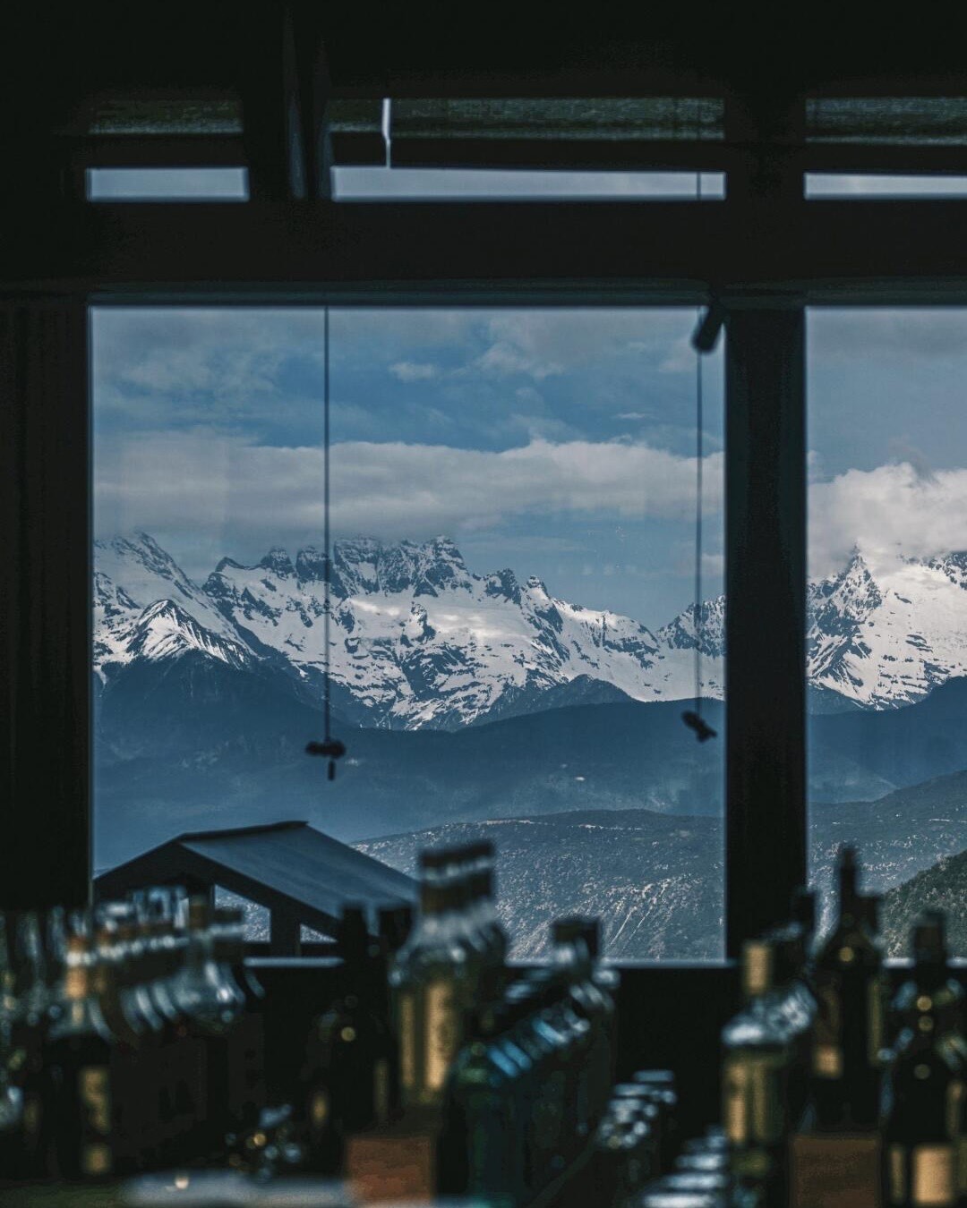 Snow-capped mountain range seen through a large lodge window above a row of bottles on a bar.