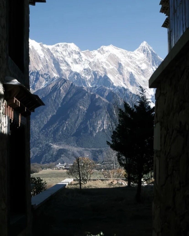 Snow-capped peaks framed between Tibetan-style stone buildings above a valley of fields and trees.