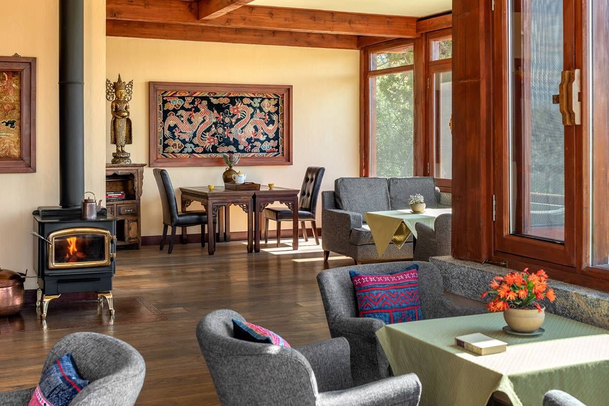 Songtsam Lodge Shangri-La — a sunlit lounge with a wood-burning stove alight, a gilded Buddha on a carved cabinet, a framed dragon carpet and armchairs by timber-framed windows