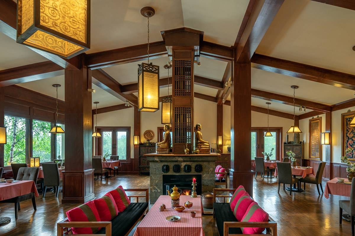 Songtsam Lodge Shangri-La — the restaurant under timber beams and lanterns, two gilded Buddhas above a central fireplace and tables laid in red check by windows onto birch trees
