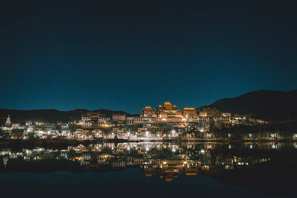 Songtsam Lodge Shangri-La — neighbouring Songzanlin Monastery lit at night under a starry sky, its gold roofs and a white stupa reflected in the lake below