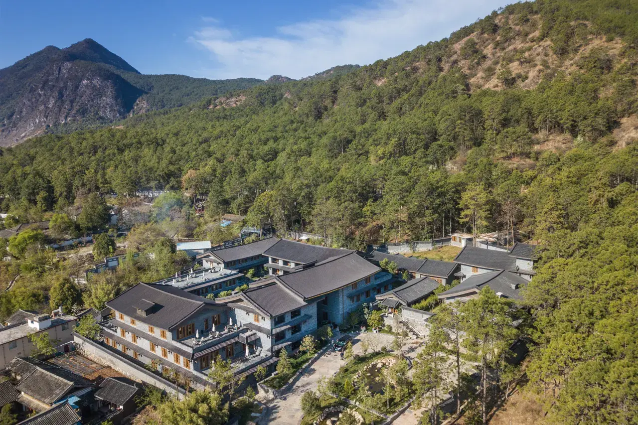 Aerial view of a grey-tiled resort complex nestled among pine forest below green and snow-dusted mountains.