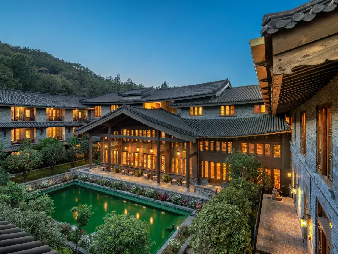 Tiered grey-tiled roof buildings surrounding a green reflecting pool and lit courtyard at dusk.