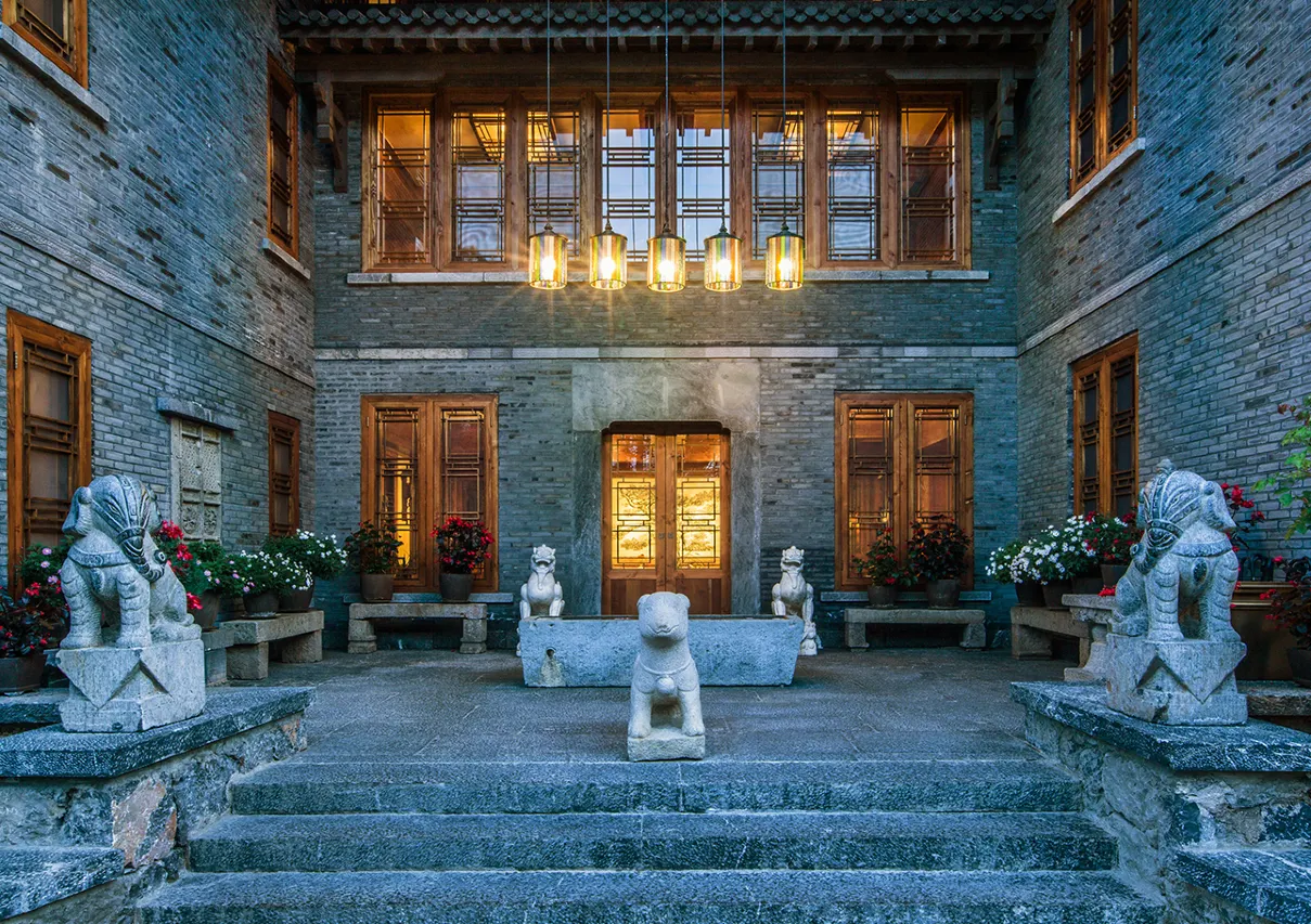 Grey-brick courtyard entrance flanked by carved stone lion statues under glowing lanterns at dusk.