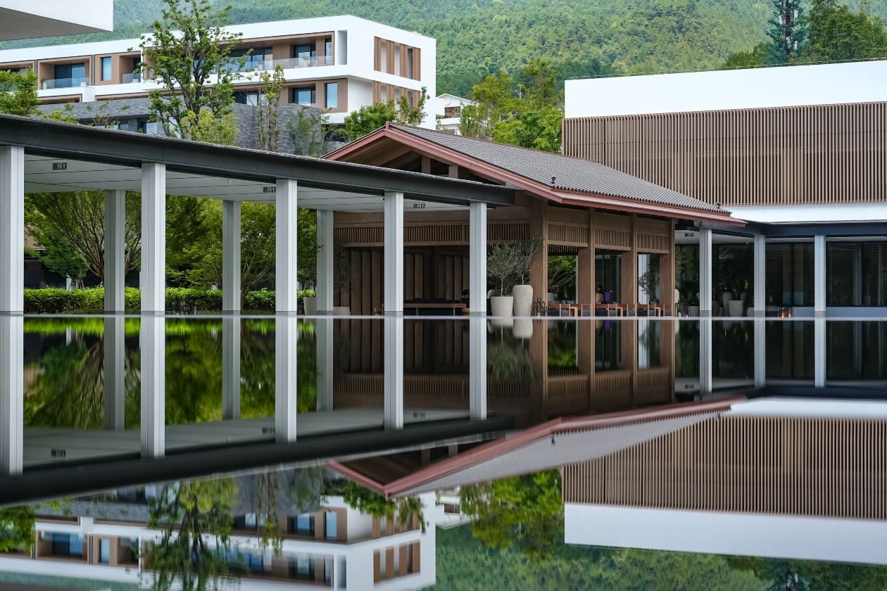 Still reflecting pool before a colonnade and a tiled-roof pavilion, white guest wings on the wooded slope above.