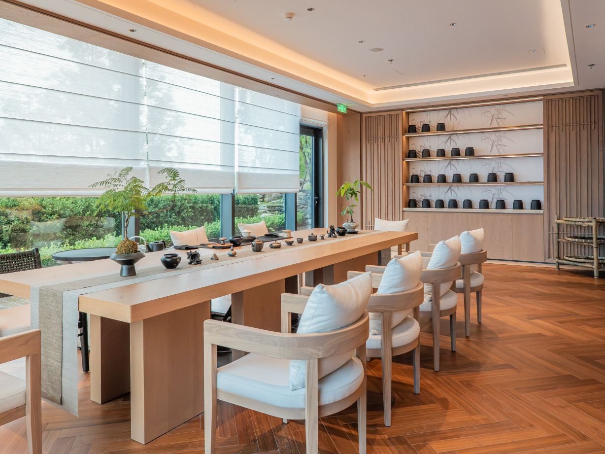 Tea room with a long pale-timber tasting table set with tea wares, shelves of teapots and a garden beyond the blinds.