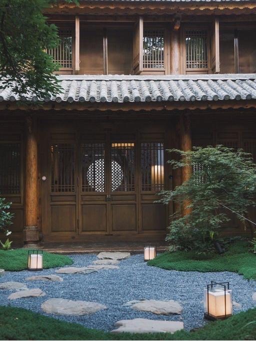Timber lodge entrance with a moss-lined gravel path, stepping stones and glowing lanterns among greenery.