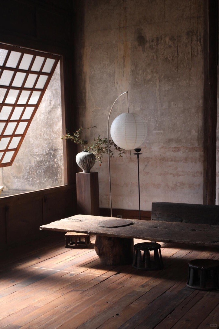 Rustic lounge with a low weathered-wood table, round paper lantern and lattice window over aged plaster walls.