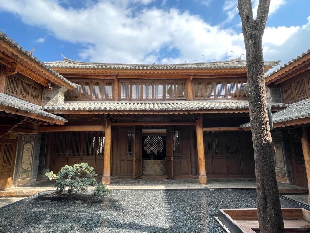 Two-storey timber courtyard building with grey tiled roofs, a reflecting pool and a bonsai tree under blue sky.