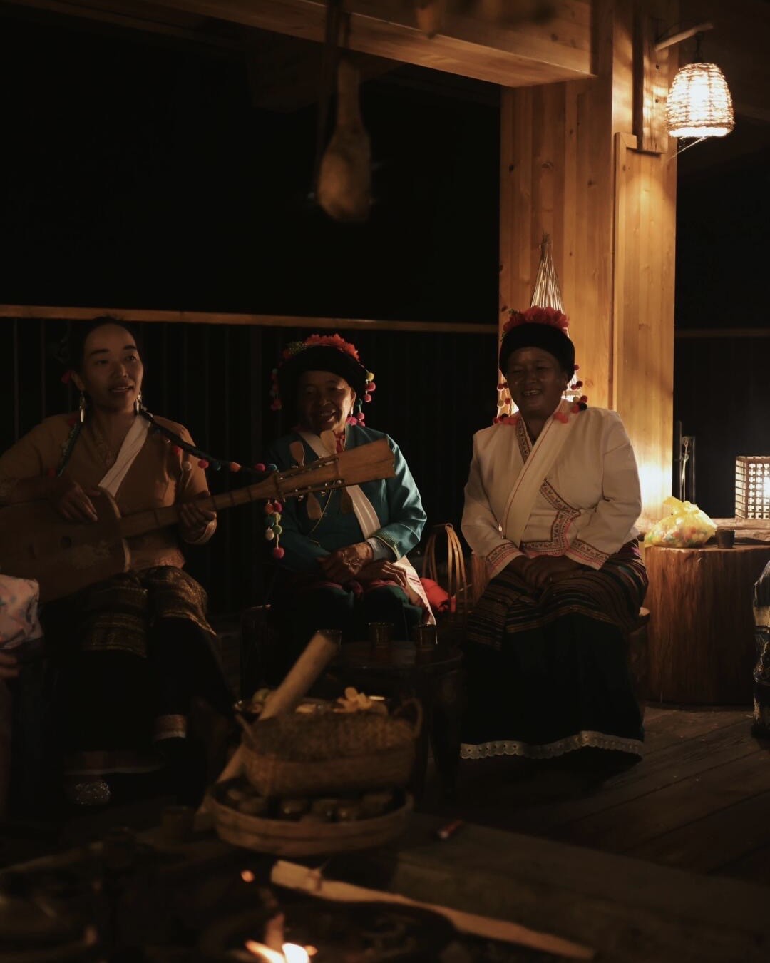 Local women in embroidered dress sing beside a fire on a timber deck as one plays a plucked lute.