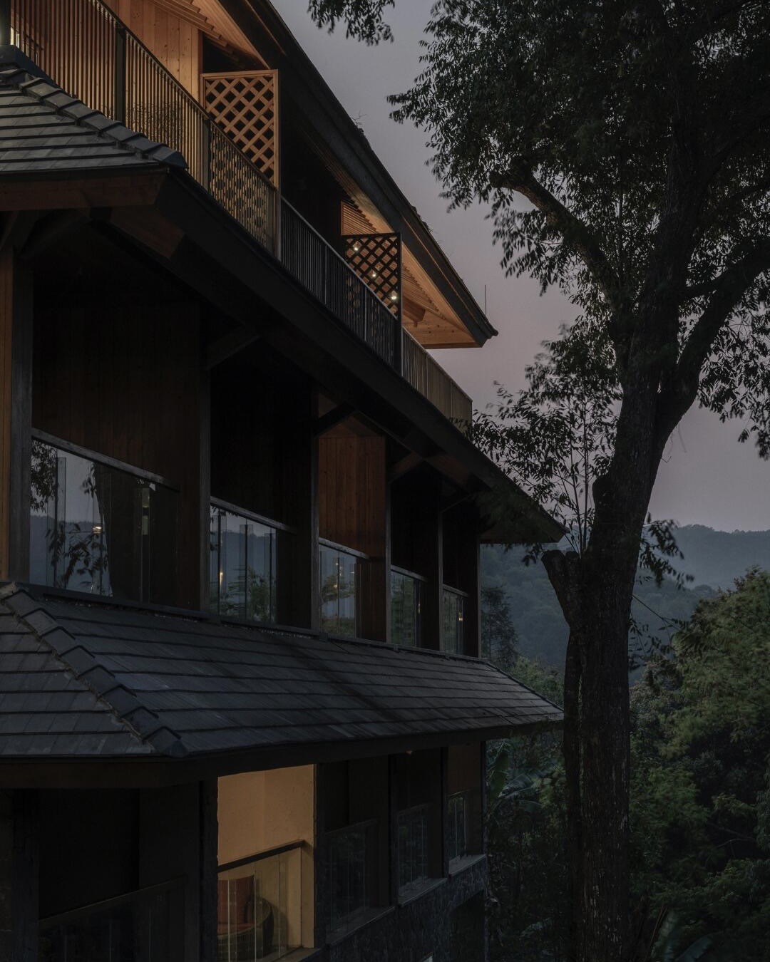 Timber lodge with tiled eaves and glass balconies at dusk, set against forested hills beside a tall tree.