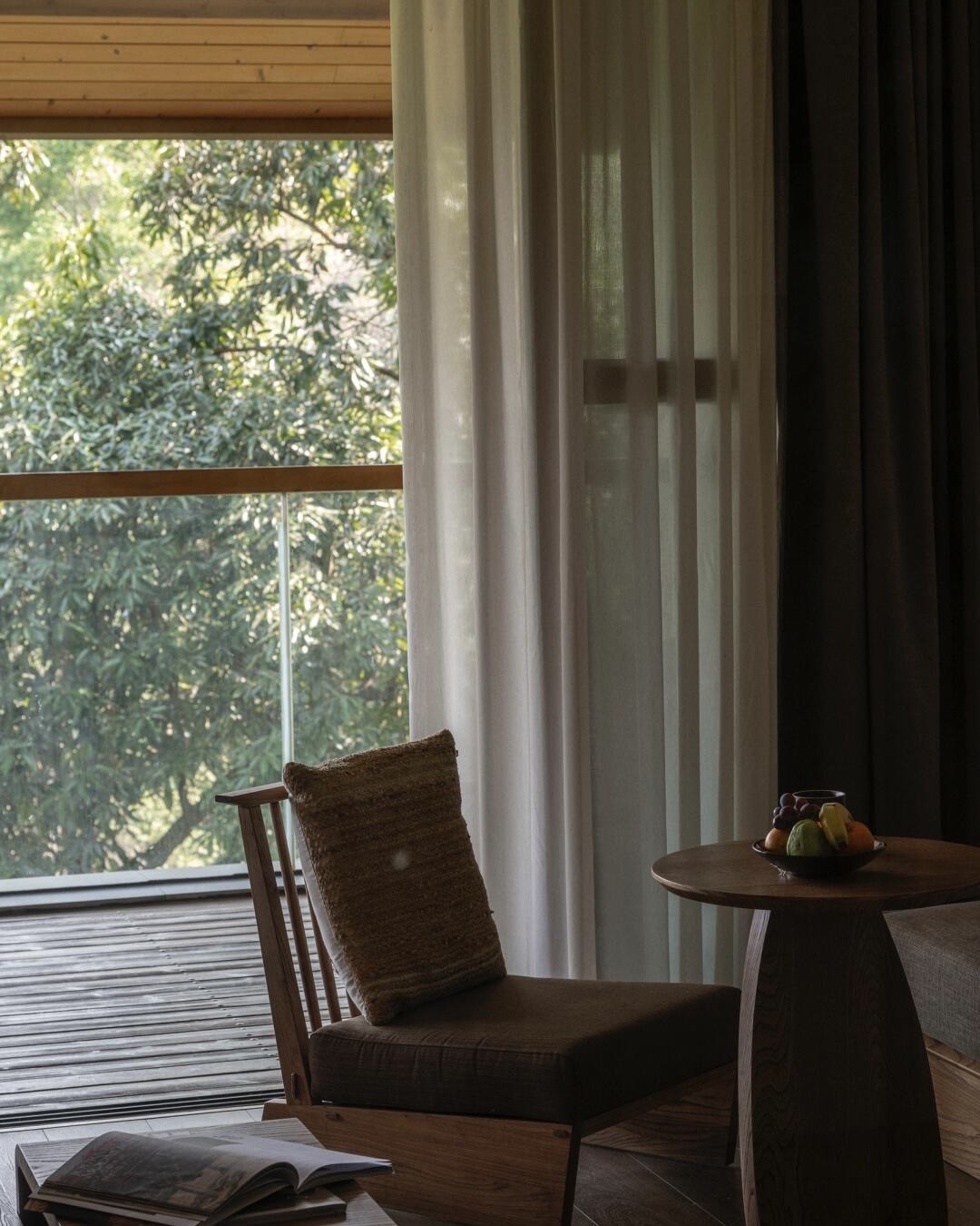 Guestroom reading nook with a wooden armchair, fruit bowl on a side table and a forest view through sheer curtains.