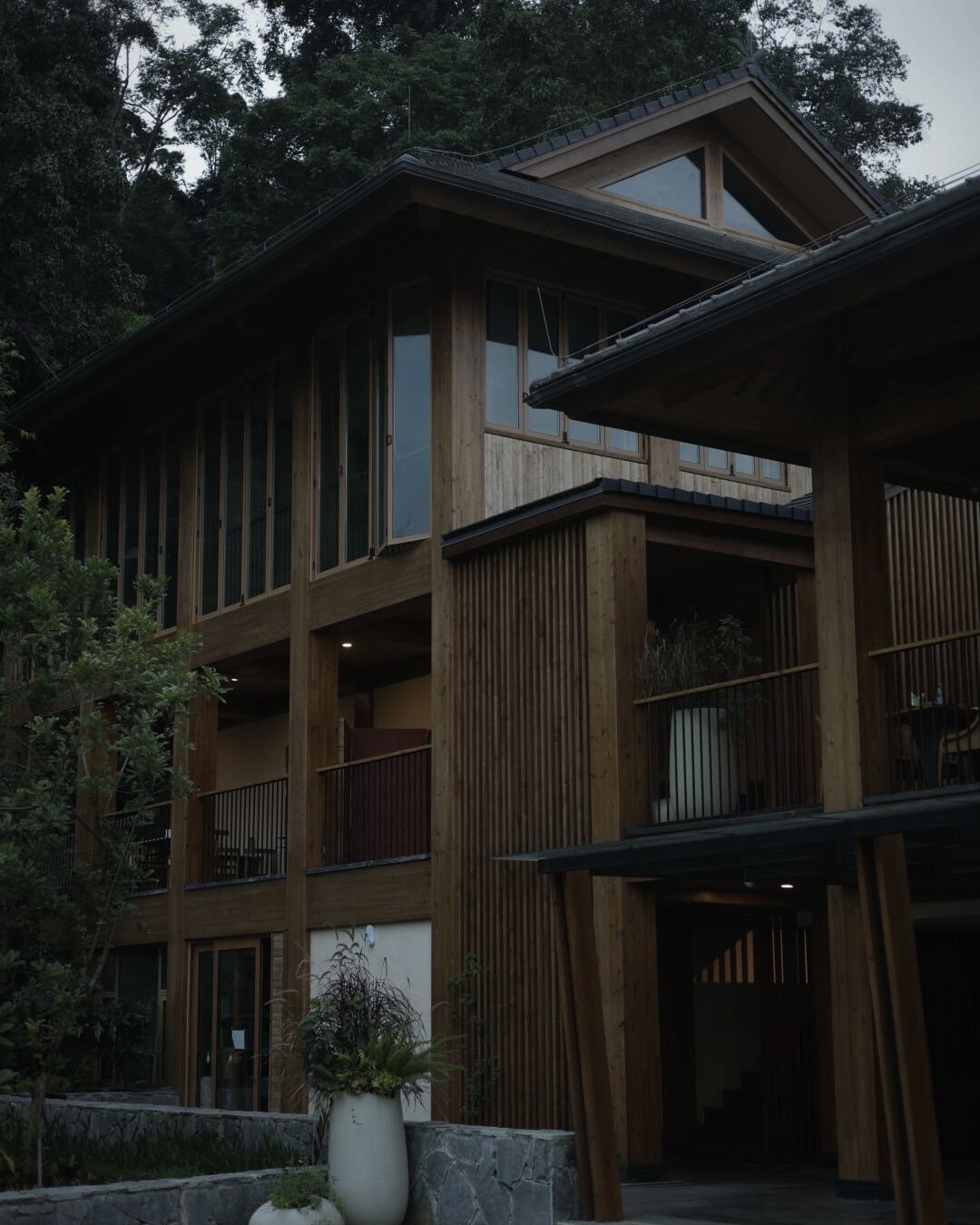 Multi-storey timber-clad lodge with slatted screens and glass balconies set against dense forest at dusk.