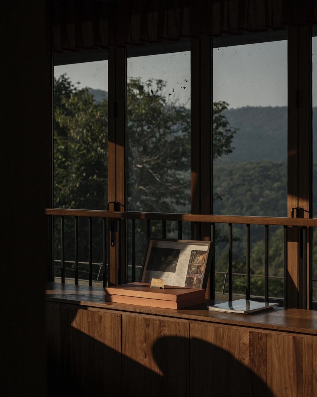 Timber window seat catching warm light, framing forested hills and folded mountains through tall panes.