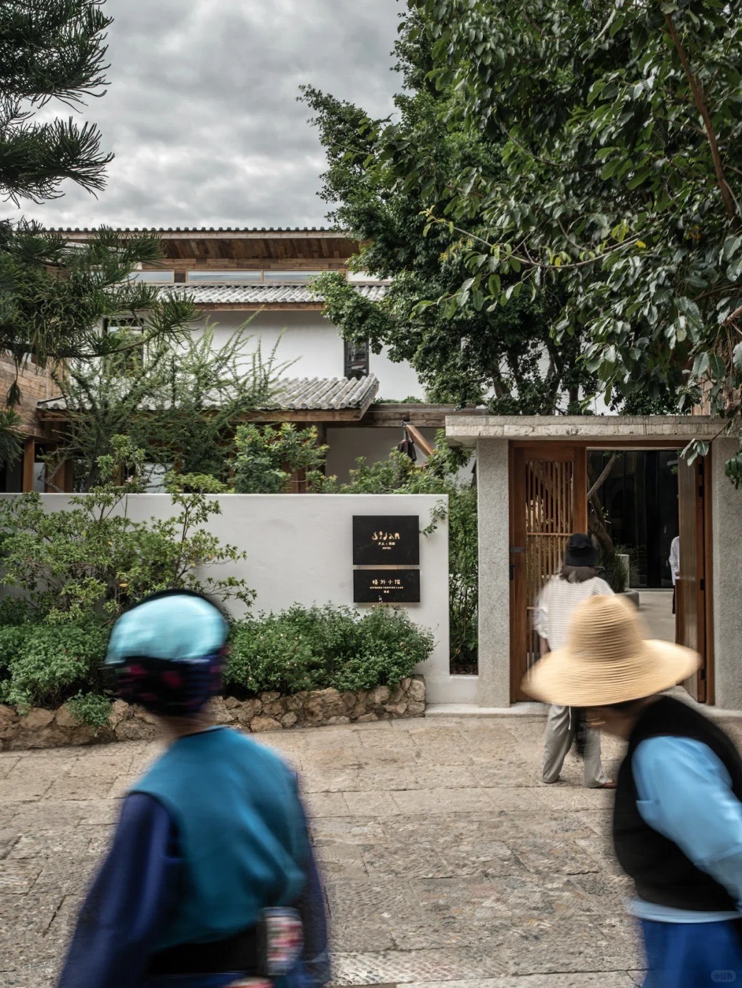 Guests in sun hats crossing a stone courtyard toward the timber gate and signed entrance of the hotel.