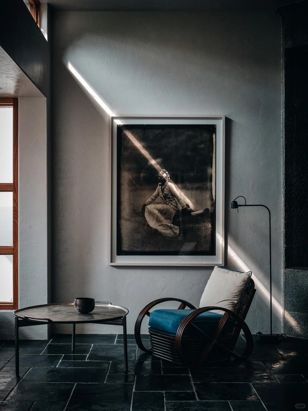 Grey plaster lounge with a framed vintage portrait, slate floor, round table and blue rattan armchair.