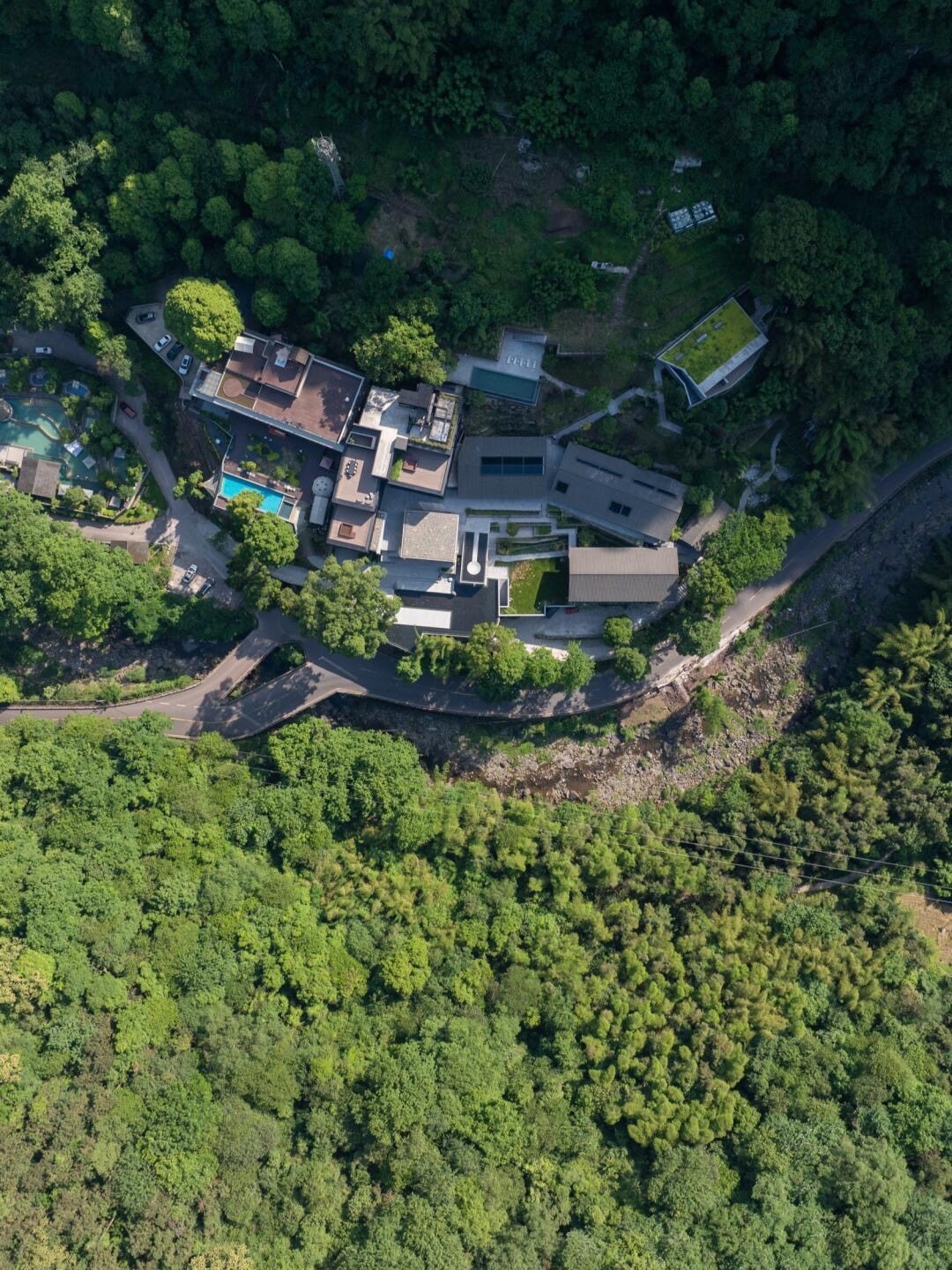 Aerial view of a dark-roofed retreat with swimming pools tucked into a steep forested hillside beside a winding road.