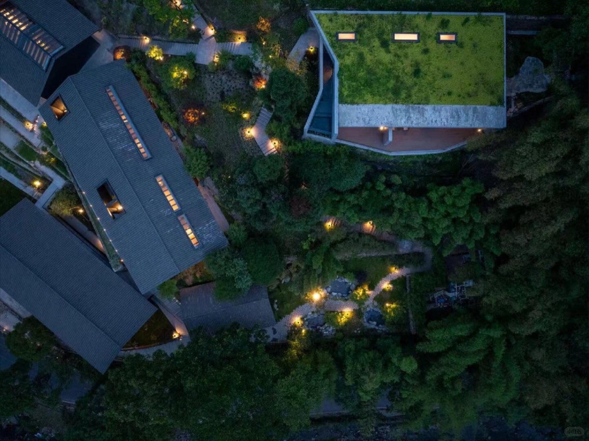 Aerial view of slate-roofed lodges and a green roof beside lit stone paths through forested gardens at night.