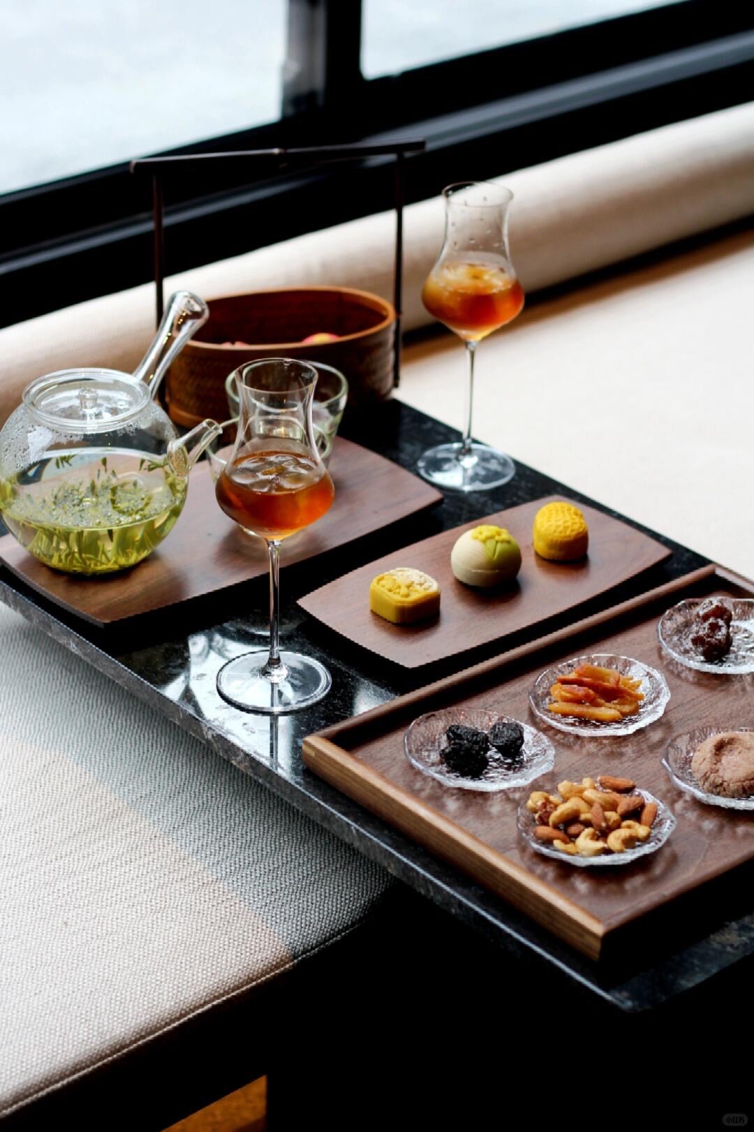 Afternoon tea set on a dark stone table with glass teapot, tea in stemmed glasses, mooncakes and dried fruits by a window.
