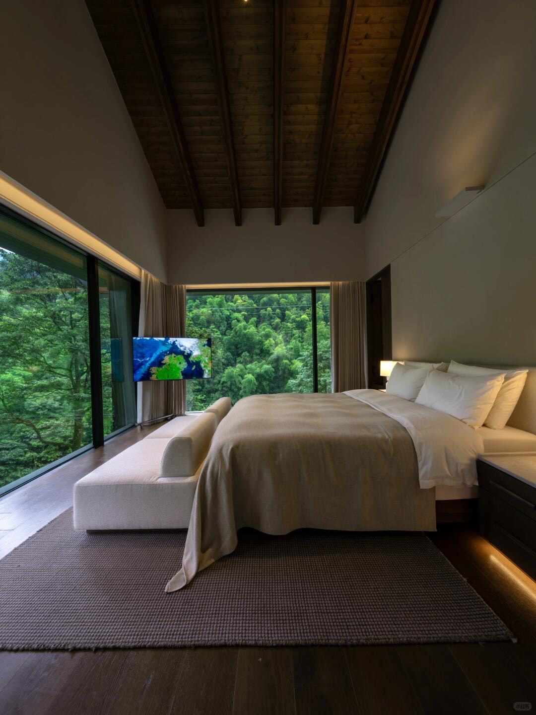 Bedroom with timber-beamed ceiling, floor-to-ceiling windows and a made bed framing a green forest view.