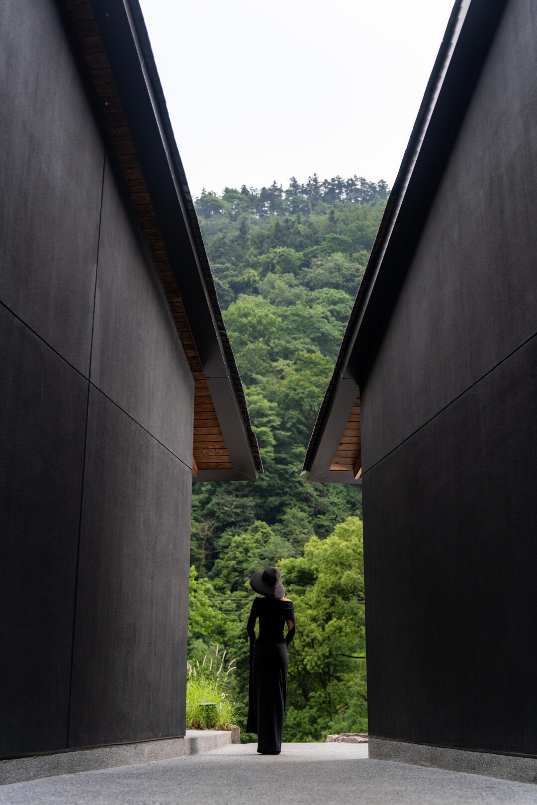 Woman in a black dress and hat standing between angular black walls that frame a green forested slope.