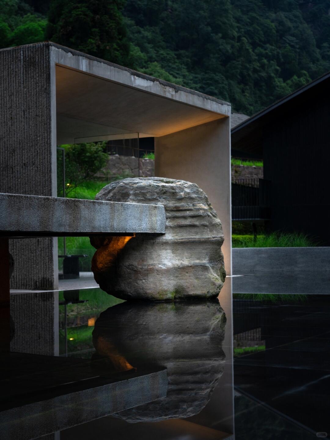 Large natural boulder set in a still reflecting pool beside a concrete pavilion, with green forested slopes behind.