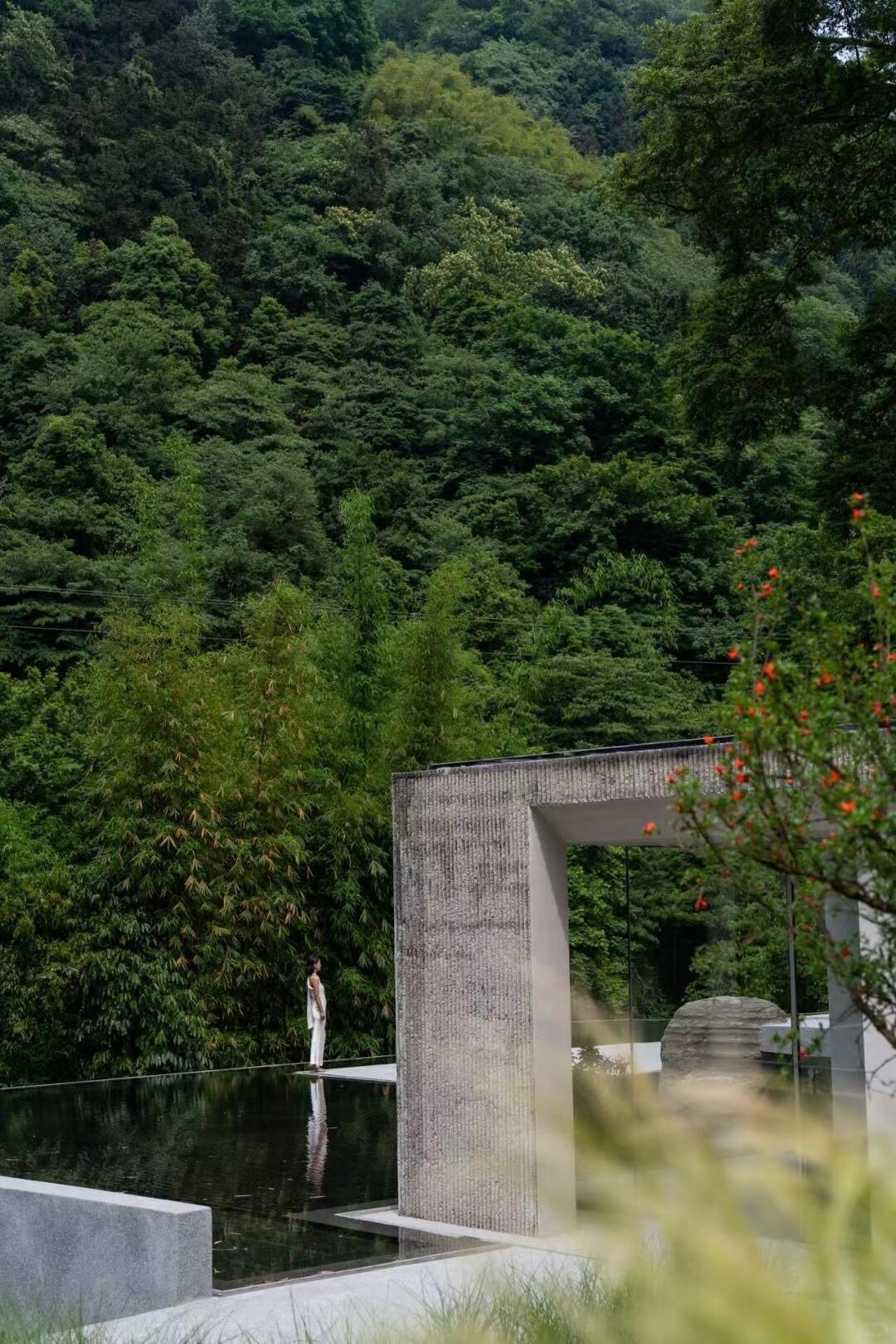 Concrete wall and reflecting pool beside dense green forest, with a woman in white at the water's edge.