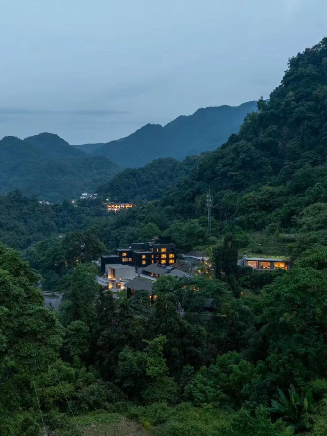 Dark modern lodge buildings glowing with warm light at dusk, nestled among dense green forest and misty mountains.