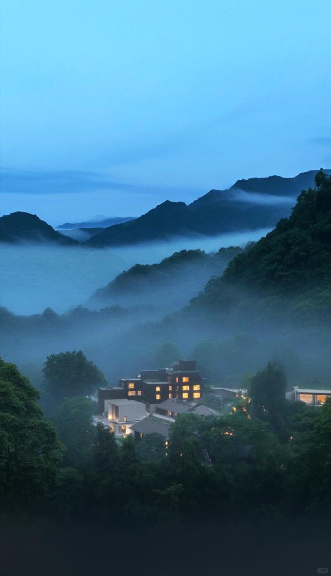 Lit hotel buildings tucked into a forested valley wrapped in low mist and layered mountains at dusk.