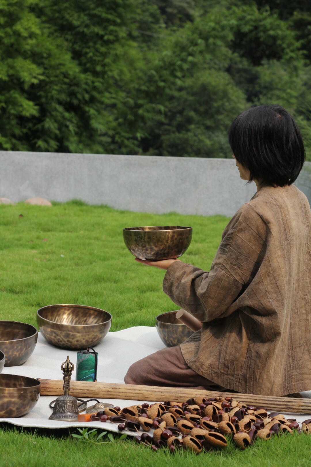 Practitioner in linen robe holding a bronze singing bowl on a lawn, surrounded by more bowls, a bell and prayer beads.